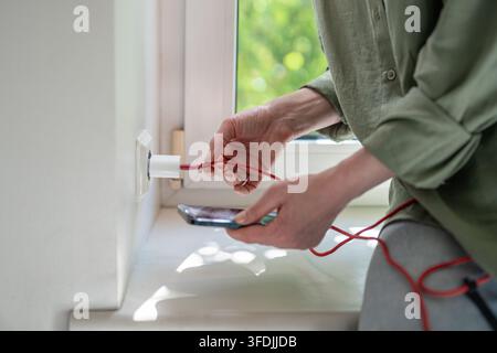 Donna regola un cavo USB rotto al caricatore da parete per mantenere la ricarica affidabile dello smartphone a casa. Foto Stock