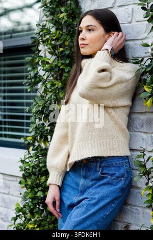 Donna adulta ispanica in maglione appoggiata a un muro di mattoni con viti, che si sistema i capelli e controlla lo smartwatch Foto Stock