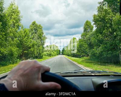 Vista attraverso il parabrezza dal sedile del conducente mostra un'autostrada a due corsie illuminata dal sole che attraversa una fitta foresta estiva sotto un cielo blu terso. Foto Stock
