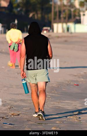 Uomo cammina a piedi nudi sulla spiaggia, indossa una felpa con cappuccio nera e pantaloncini. Porta una bottiglia d'acqua blu, telefono in mano. La luce del sole proietta lunghe ombre sulla sabbia. Ba Foto Stock
