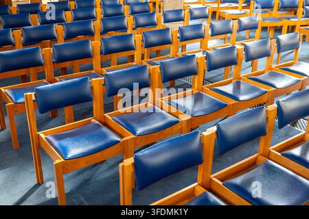 Una vista ad angolo interno di sedie di legno vuote con sedili e schienali in pelle blu, disposte a file su un tappeto blu. Foto Stock