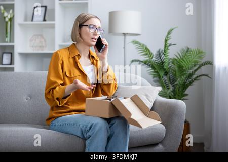 Donna arrabbiata sul divano che parla al telefono, lamentandosi di un pacco errato o danneggiato dagli acquisti online mentre tiene in mano una scatola di cartone aperta a casa, frustrata e in cerca di aiuto Foto Stock