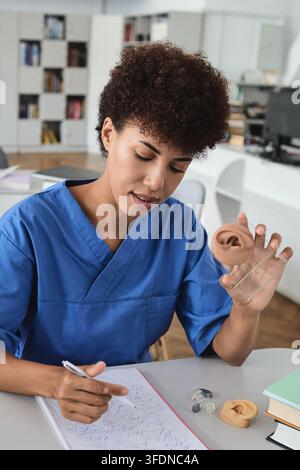 Primo piano verticale di uno studente di medicina afroamericano che tiene un modello anatomico dell'orecchio mentre prende appunti. Audiologia clinica e diagnostica uditiva Foto Stock
