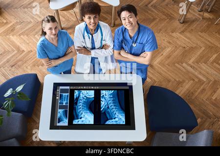 Ritratto aereo di un team clinico diversificato e sicuro di sé in piedi accanto a un tavolo di diagnostica digitale che visualizza radiografie delle vertebre cervicali Foto Stock