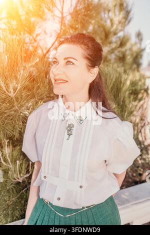 Ritratto di donna nel bosco, donna sorridente in camicetta vintage a fiori in piedi nella luce dorata tra i pini per un concetto di stile di vita Foto Stock