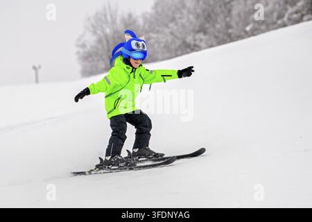 Giovane sciatore con una giacca neon brillante e un casco a carattere divertente, che scia lungo una pendice innevata. Sport invernali emozionanti e avventura all'aperto Foto Stock