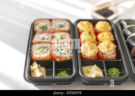 Assortimento di rollini di sushi freschi in scatola nera da asporto con rollini al salmone, avocado, formaggio cremoso e salsa, serviti con wasabi e zenzero su sfondo bianco Foto Stock