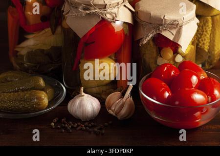Su un tavolo di legno ci sono barattoli di vetro con verdure sottaceto, una ciotola con pomodorini, cetrioli sottaceto, teste d'aglio intere e grani di pepe - un ru Foto Stock