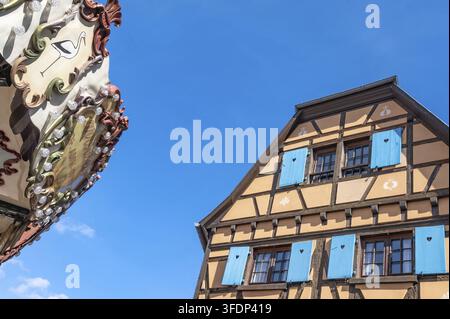 Gabbia di una tradizionale casa a graticcio con persiane azzurre accanto al tetto di una giostra nostalgica a tema cicogna sotto un cielo blu, Obernai, B Foto Stock