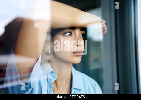 Giovane donna che guarda pensierosa fuori da una finestra con un'espressione triste, riflettendo su emozioni, solitudine e isolamento all'interno Foto Stock