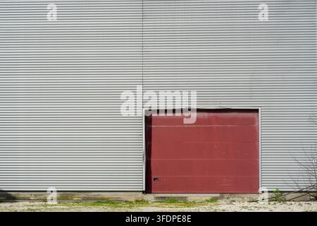Esterno industriale minimale con una porta del garage rossa chiusa inserita in una parete di magazzino in lamiera ondulata grigia, linee orizzontali e texture consumata c Foto Stock