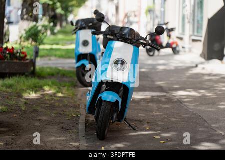 Due monopattini elettrici blu in condivisione sono parcheggiati su una striscia di parcheggio segnalata accanto a un marciapiede alberato, le flotte di noleggio occupano lo spazio del marciapiede che un tempo era riservato alle auto Foto Stock