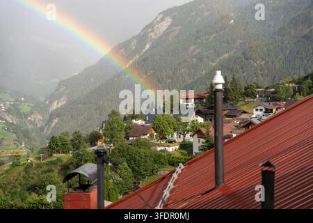 Un arcobaleno luminoso si inarca su un villaggio alpino con tetti rossi, alberi verdi lussureggianti e uno sfondo montano boscoso, che trasmette calma, freschezza e il Foto Stock