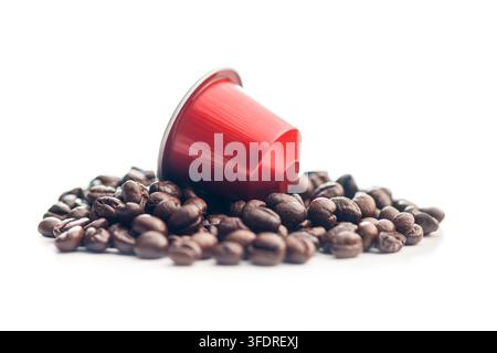 Una capsula di caffè rossa monodose appoggiata su un mucchio di chicchi di caffè tostati su uno sfondo bianco pulito, che suggerisce un ricco sapore di espresso, mattina Foto Stock