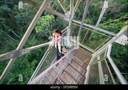 Donna avventurosa in piedi su una torre della canopia in metallo sopra una fitta foresta pluviale, con uno zaino e un binocolo, sorride e guarda verso l'alto mentre esplora la chioma Foto Stock