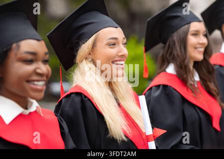 Giovane donna bionda e carina sorridente in mezzo a un gruppo internazionale di studenti, indossa costumi da laurea e tiene diplomi, laureata multirazziale Foto Stock