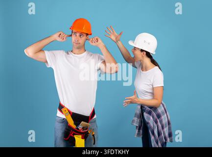 Coppia in hardhats che discutono durante la ristrutturazione, l'uomo irritato tappare le orecchie con le dita Foto Stock