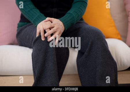 Anziana coreana che tiene il ginocchio dolorante mentre è seduta sul divano a casa Foto Stock