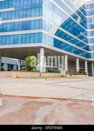 Edificio moderno in vetro si erge alto ed elegante. Le finestre colorate di blu riflettono il cielo e l'ambiente circostante. Piazza in cemento conduce all'ingresso con pilastri. Gree Foto Stock