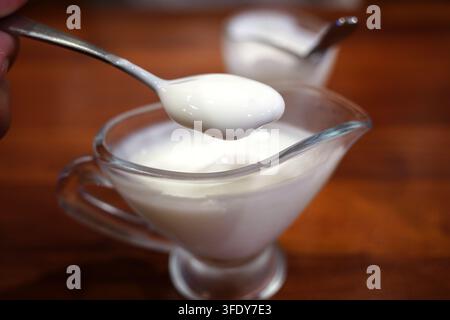 Yogur bianco e cremoso su un cucchiaio tenuto sopra un piccolo contenitore di vetro per salse Foto Stock