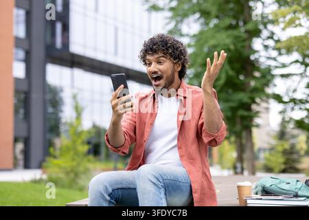 Giovane studente felice che mostra una reazione sorpresa ed entusiasta mentre guarda il suo telefono cellulare su una panchina del campus universitario, ricevendo buone notizie o vincendo online Foto Stock
