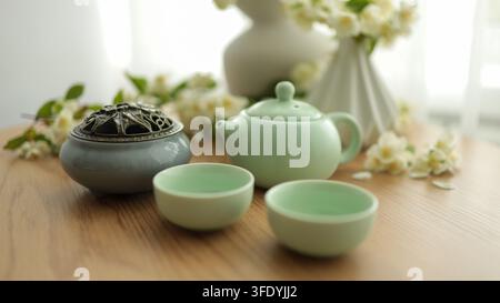 Servizio da tè verde tradizionale con teiera in ceramica, piccole tazze e vaso con fiori bianchi su tavolo di legno, composizione pacifica di natura morta. Foto Stock