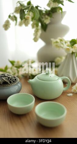 Servizio da tè verde tradizionale con teiera in ceramica, piccole tazze e vaso con fiori bianchi su tavolo di legno, composizione pacifica di natura morta. Foto Stock