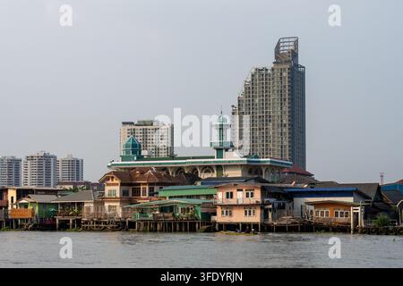 NONTHABURI, THAILANDIA — 20 GENNAIO 2026: La moschea Ta-It e le case di legno sul lungofiume si ergono davanti ai moderni grattacieli residenziali sul fiume Chao Phraya. Foto Stock