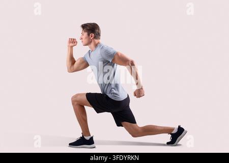 Vista laterale di un forte ragazzo millenario in abbigliamento sportivo che fa affondo su sfondo studio chiaro Foto Stock