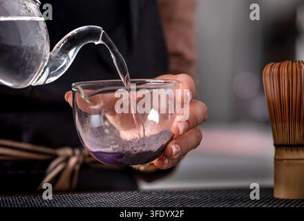 Barista femminile che versa acqua calda nella polvere di tè matcha blu, mentre il processo mostra un primo piano di una mano Foto Stock