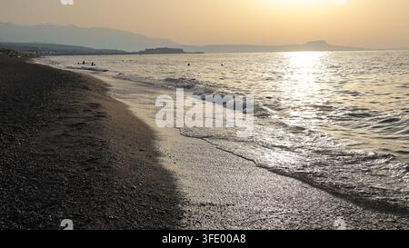 Vista verso ovest lungo la spiaggia sabbiosa di Rethymno con il sole basso che splende sul mare a Creta, Grecia Foto Stock