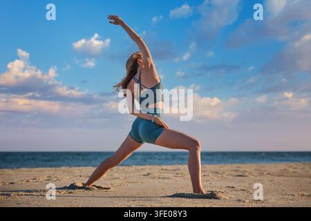 Sulla sabbia della spiaggia, una donna tiene una variazione di yoga Viparita Virabhadrasana semi-legata, Reverse Warrior, con un braccio sopra la testa e l'altro dietro la schiena Foto Stock