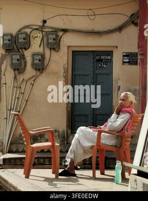 VARANASI, INDIA - 23 gennaio 2026: Un uomo anziano con una kurta bianca e una sciarpa rosa è seduto a riposare su una sedia di plastica su una veranda residenziale Foto Stock