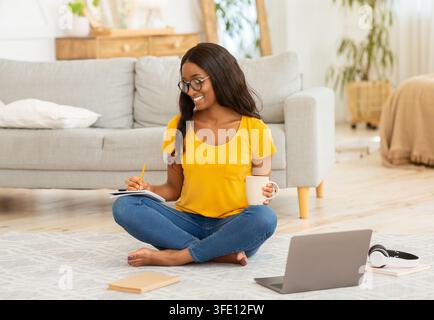 Concetto di formazione a distanza. Bella studentessa nera che scrive qualcosa nel notebook di fronte al portatile a casa Foto Stock