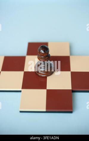 Primo piano di un pedone di legno scuro su una piccola croce di scacchi rosso e panna, su uno sfondo blu pallido Foto Stock