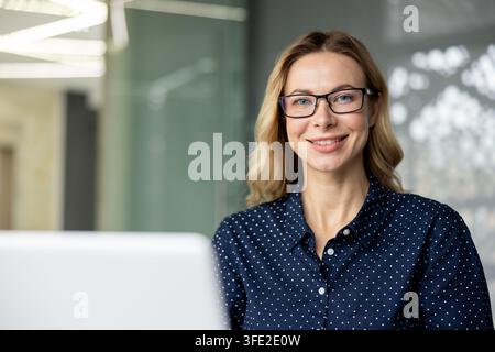 Giovane donna d'affari professionista sorridente, con occhiali e che guarda la fotocamera mentre è seduta alla sua scrivania, simboleggiando successo, sicurezza di sé e crescita professionale in un ambiente d'ufficio moderno Foto Stock