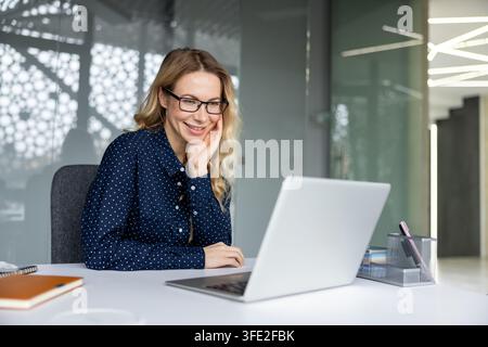 Donna d'affari sorridente con occhiali che collabora da remoto e interagisce con contenuti digitali sul suo laptop, incarnando un ambiente di lavoro professionale e positivo mentre utilizza la tecnologia Foto Stock