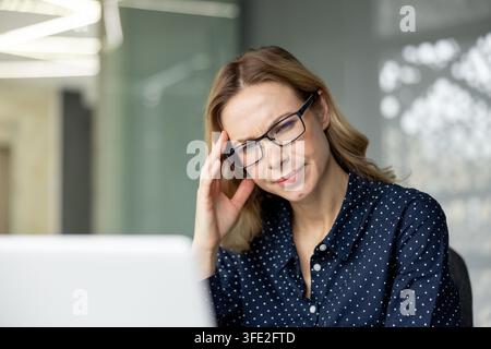 Donna con occhiali che soffre di un doloroso mal di testa mentre lavora da remoto sul suo laptop in un ufficio moderno, si sente stanca, frustrata e stressata con sintomi di burnout Foto Stock