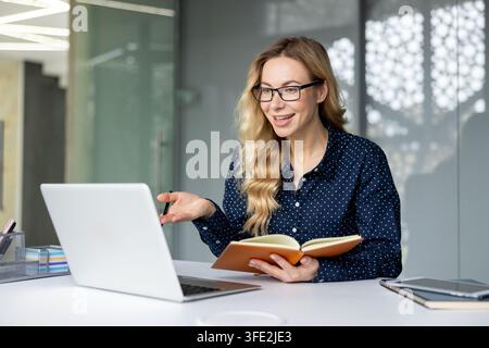 Donna con occhiali e una camicia blu a pois che lavora da un ufficio moderno, partecipa attivamente a una riunione virtuale mentre tiene un quaderno e gesticola con la mano Foto Stock