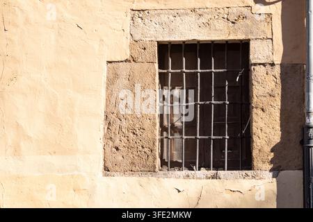 Finestra con sbarre di ferro illuminata dal sole, incorniciata da pietra ruvida e intonaco sbiadito su un edificio storico a León, Spagna, enfatizzando la trama, l'età e il caldo Mediterraneo Foto Stock