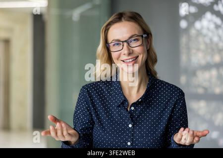 Donna d'affari bionda con occhiali e camicia a pois, guarda la telecamera con un sorriso accogliente e mani aperte, comunica sicurezza e cordialità in un ambiente d'ufficio Foto Stock