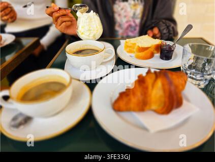 Prima piano di una colazione francese tradizionale su un tavolo da caffè con croissant fresco, tazze di caffè, panna montata, pane e marmellata. Foto Stock