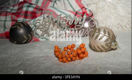 Insegna argentata scintillante con auguri di Buon Natale circondata da eleganti palline dell’albero e bacche di sorbo arancioni brillanti su una coperta di peluche bianca, morbido tartan a quadri rossi Foto Stock