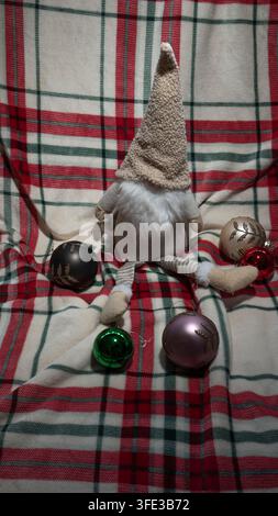 Simpatico giocattolo di gnomo di Natale in peluche con cappello alto seduto su una coperta scozzese rossa circondato da palline verdi, rosa, rosse, nere e crema, verticale fe Foto Stock