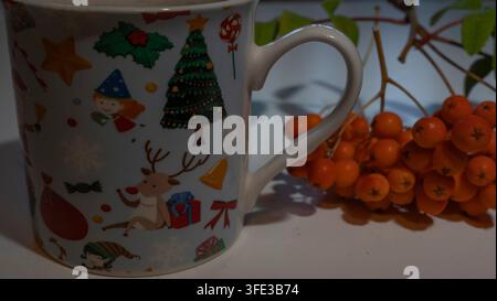 Prima vista laterale di una tazza di ceramica bianca festosa decorata con illustrazioni natalizie colorate con renne, albero di Natale ed elfo, accanto a un'arancia Foto Stock