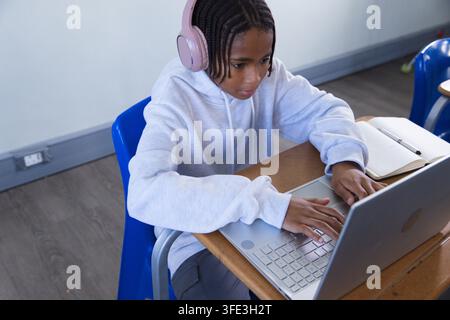 Adolescente afroamericano che digita su un laptop argentato alla scrivania di scuola indossando una felpa con cappuccio grigia e un auricolare rosa Foto Stock