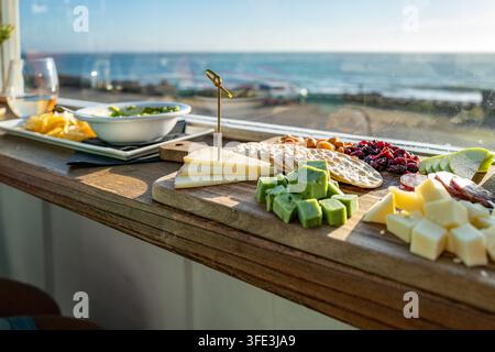 Tavola di salumi gourmet con formaggi assortiti e cracker servita su una ringhiera del balcone che si affaccia sulla costa dell'Oceano Pacifico. Foto Stock