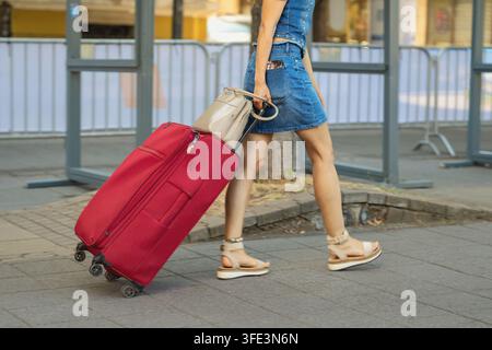 Donna che tira una valigia rossa in città. Donna che cammina all'aperto mentre tira una grande valigia rossa con le ruote e porta una borsa a mano. Stile di vita da viaggio, turismo Foto Stock