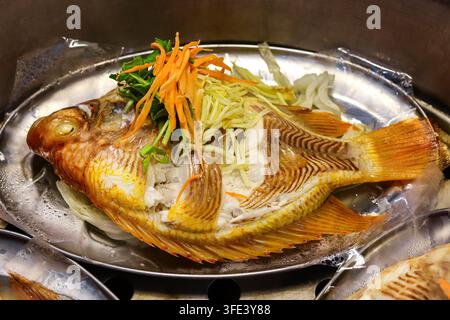 Tilapia rosso intero, un pesce d'acqua dolce, cotto a vapore con salsa di soia e zenzero grattugiato, servito con verdure su un vassoio in acciaio inossidabile in una trattoria. Foto Stock