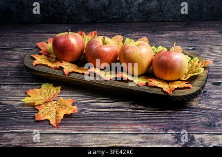 Quattro mele Gala rosse e gialle disposte su foglie autunnali colorate all'interno di un vassoio di legno rustico scuro, con foglie extra su un tavolo di legno. Foto Stock